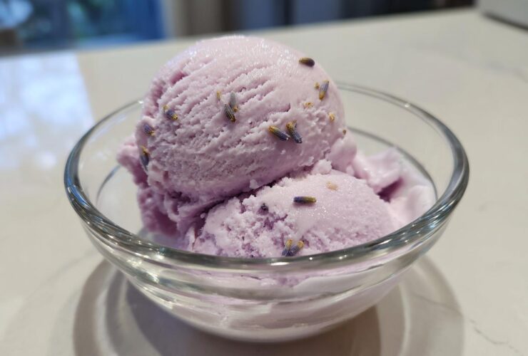 Two scoops of creamy purple Lavender Honey Ice Cream garnished with dried lavender buds in a clear glass bowl.