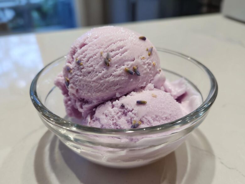 Two scoops of creamy purple Lavender Honey Ice Cream garnished with dried lavender buds in a clear glass bowl.