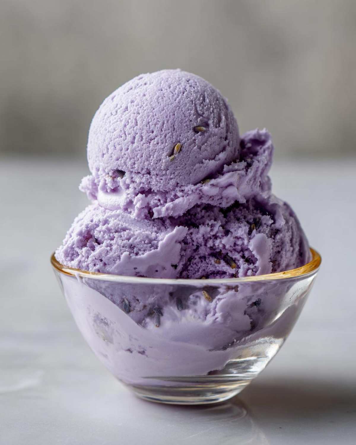Two scoops of creamy purple Lavender Honey Ice Cream served in a clear glass bowl, with visible lavender buds.