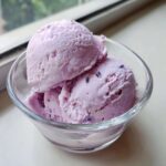 Two scoops of creamy purple Lavender Honey Ice Cream in a glass bowl, garnished with dried lavender buds.