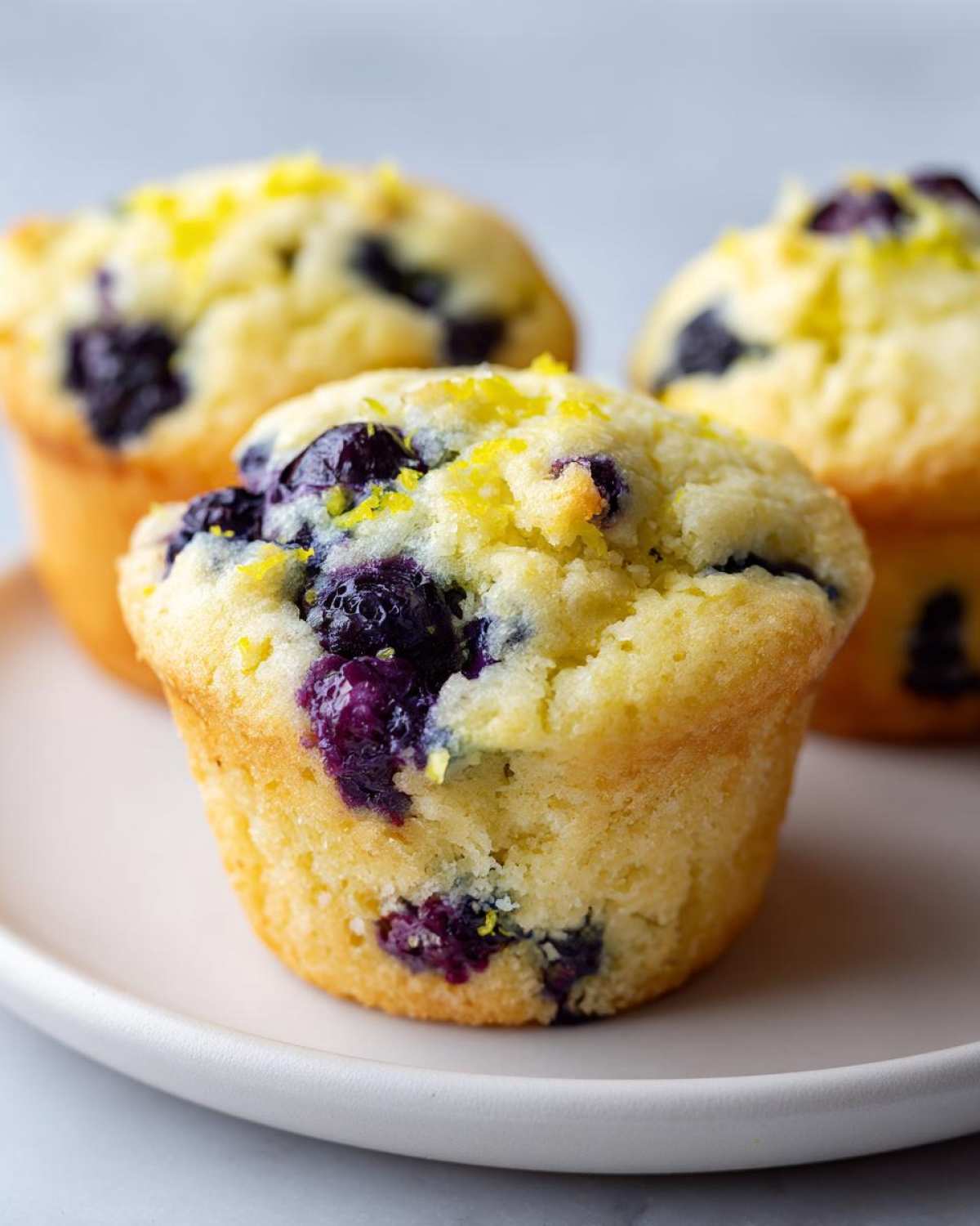 Close-up of three perfect lemon blueberry muffins topped with fresh blueberries and lemon zest.