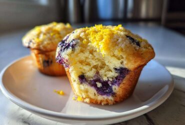 Close-up of a perfect lemon blueberry muffin, split open to reveal blueberries and topped with lemon zest.