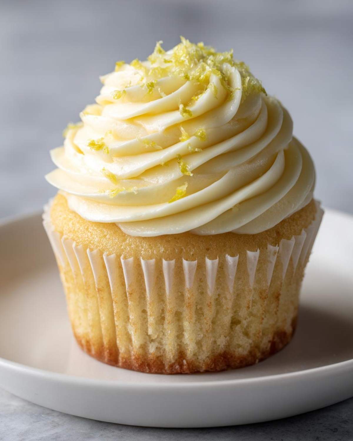 A single, perfectly frosted Lemon Vanilla Cupcake topped with fresh lemon zest on a white plate.