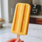 A hand holding a bright orange Mango Lassi Popsicle with vertical ridges.