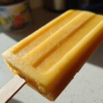 Close-up of a bright orange Mango Lassi Popsicle on a wooden stick, showing its textured surface.