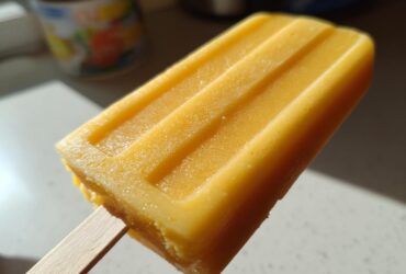 Close-up of a bright orange Mango Lassi Popsicle on a wooden stick, showing its textured surface.