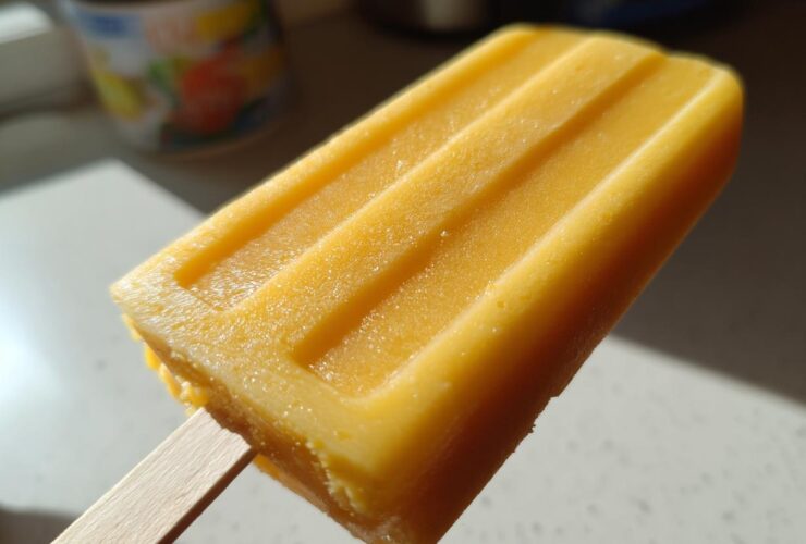 Close-up of a bright orange Mango Lassi Popsicle on a wooden stick, showing its textured surface.