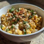 A close-up of a white bowl filled with creamy Mexican street corn salad, topped with crumbled cheese, cilantro, and chili powder.
