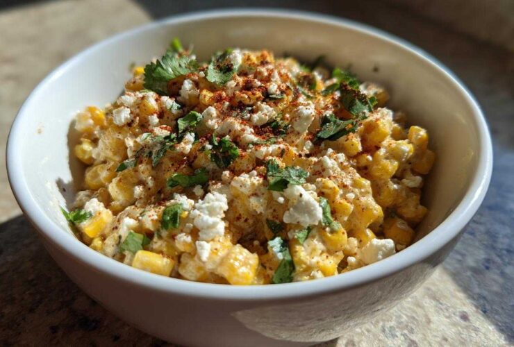 A close-up of a white bowl filled with creamy Mexican street corn salad, topped with crumbled cheese, cilantro, and chili powder.