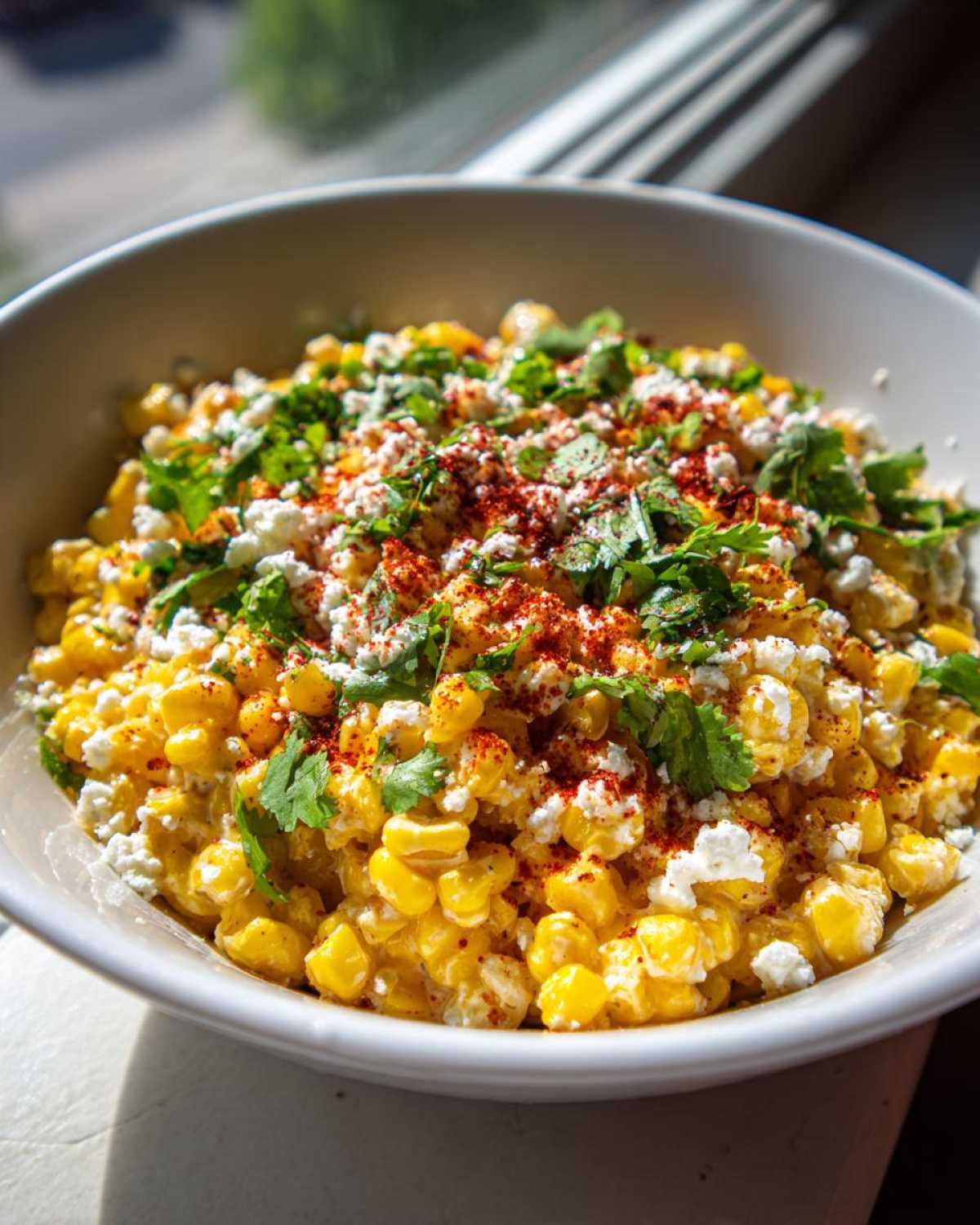 A close-up of a bowl of Mexican street corn salad, featuring corn, crumbled cheese, cilantro, and chili powder.