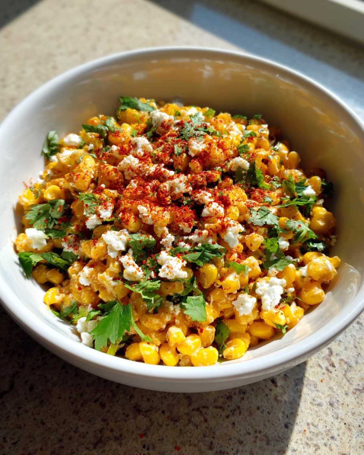 A close-up of Mexican street corn salad in a white bowl, topped with crumbled cheese, cilantro, and chili powder.