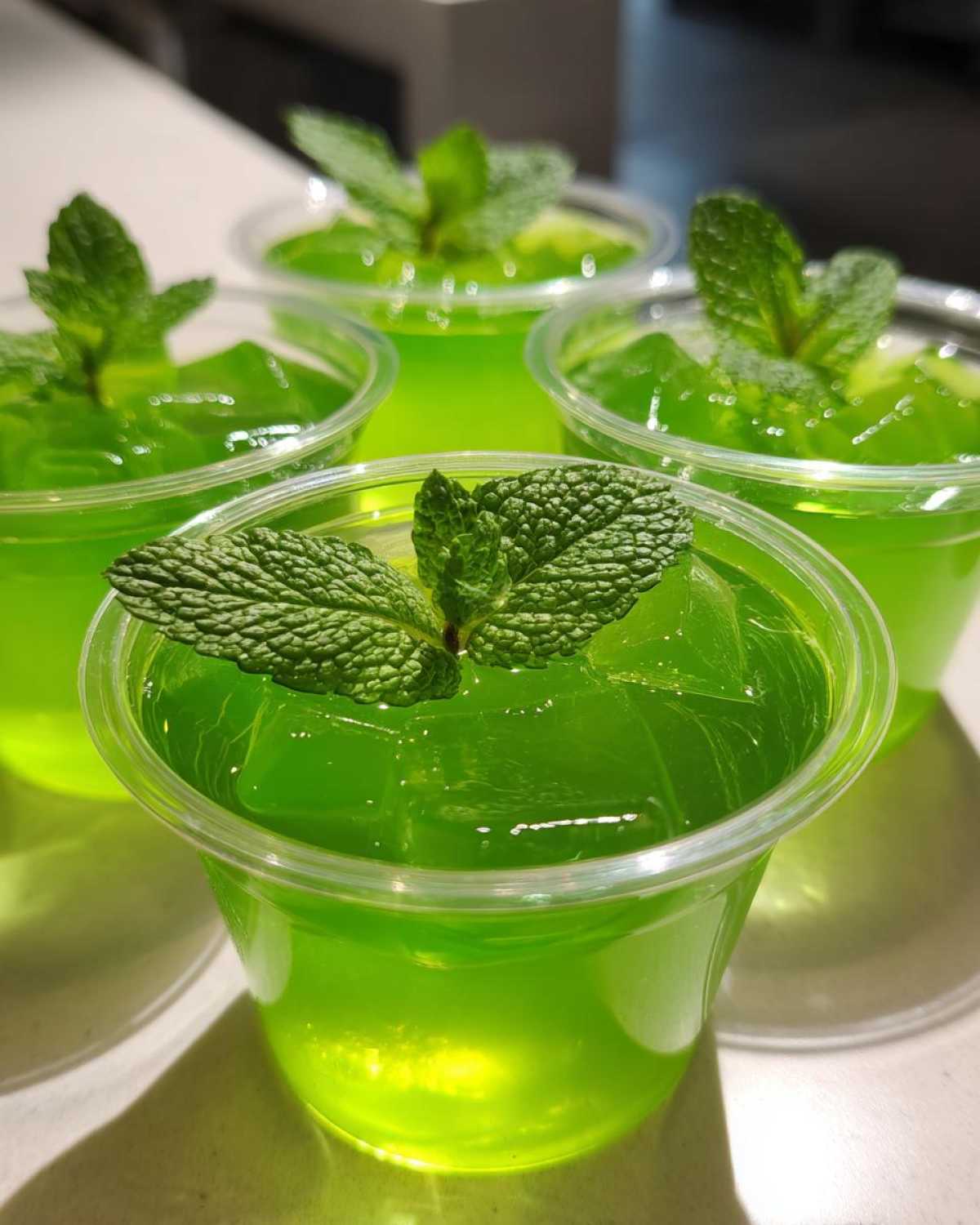 Close-up of four Mint Julep Jello Shots in clear plastic cups, each topped with fresh mint leaves and ice.