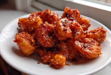 A close-up of a plate piled high with glistening Nashville Hot Shrimp, coated in a fiery red sauce and sprinkled with spices.