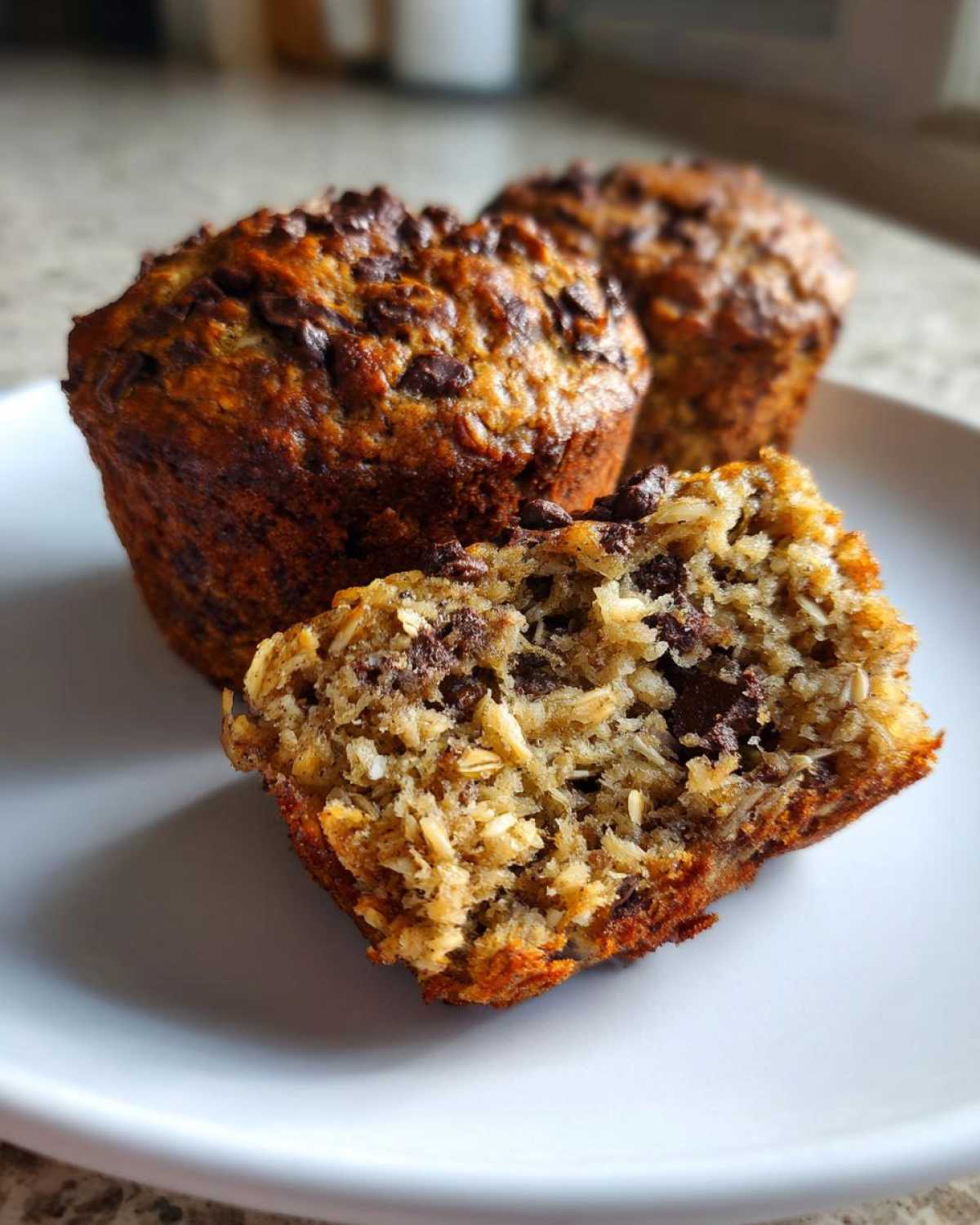Close-up of a halved oatmeal chocolate chip muffin on a white plate, revealing oats and chocolate chips inside.