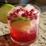 A close-up of a Pomegranate Guava Mocktail garnished with fresh lime and pomegranate seeds, filled with ice and bubbly liquid.