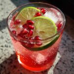 A close-up of a Pomegranate Guava Mocktail in a glass, garnished with lime slices and pomegranate seeds, with ice and bubbles.