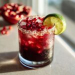 A close-up of a Pomegranate Margarita cocktail, garnished with fresh pomegranate seeds and a lime wedge, with a salt-rimmed glass.