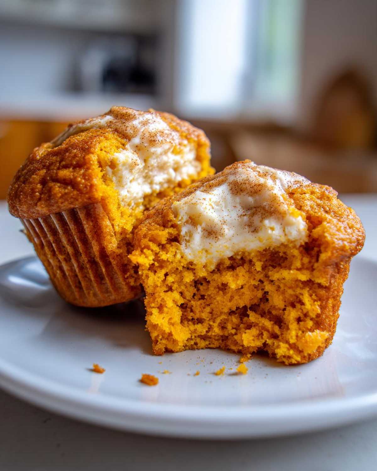 Close-up of two Pumpkin Cream Cheese Muffins on a white plate, one cut in half revealing a creamy center and sprinkled with cinnamon.