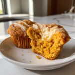 Close-up of two Pumpkin Cream Cheese Muffins on a white plate, one is cut in half revealing the cream cheese filling.