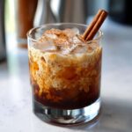 A close-up of a Pumpkin Spice Cold Brew Cocktail with cream, ice, cinnamon, and a cinnamon stick.