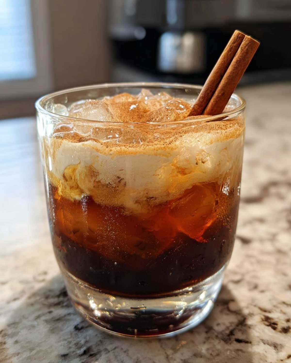 A refreshing Pumpkin Spice Cold Brew Cocktail in a glass, topped with cream, cinnamon, and a cinnamon stick.