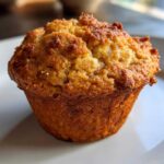 A close-up, golden-brown pumpkin spice muffin sits on a white plate, showcasing its textured top.
