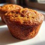 Close-up of two golden-brown pumpkin spice muffins on a white plate, bathed in warm sunlight.