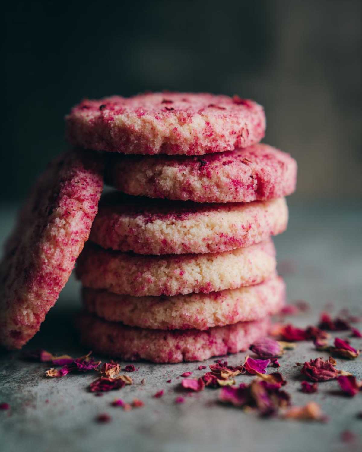 A stack of pink-flecked Raspberry Rosewater Shortbread cookies with dried rose petals scattered around.