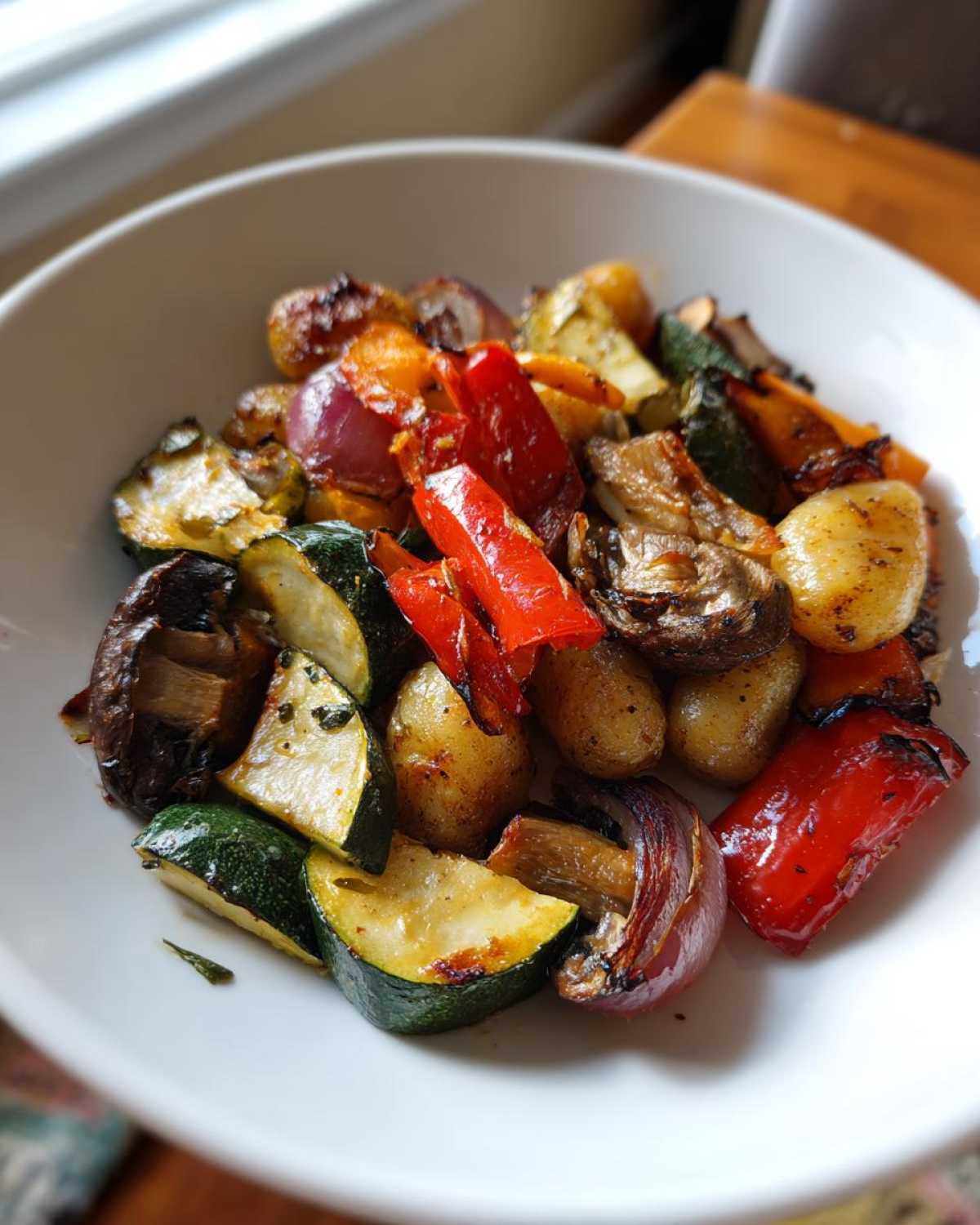 A close-up of a white bowl filled with colorful roasted vegetables and gnocchi, featuring zucchini, red bell peppers, mushrooms, red onion, and potatoes.