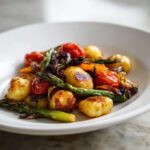 A close-up of a white plate filled with golden-brown sheet pan gnocchi and roasted vegetables including asparagus and cherry tomatoes.