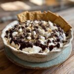 A close-up of a S'mores Cheesecake Dip topped with mini marshmallows, chocolate chips, and graham cracker crumbs, served with graham crackers.