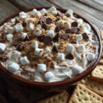 A close-up of a S'mores Cheesecake Dip in a brown dish, topped with marshmallows, chocolate chips, and graham cracker crumbs, with graham crackers on the side.