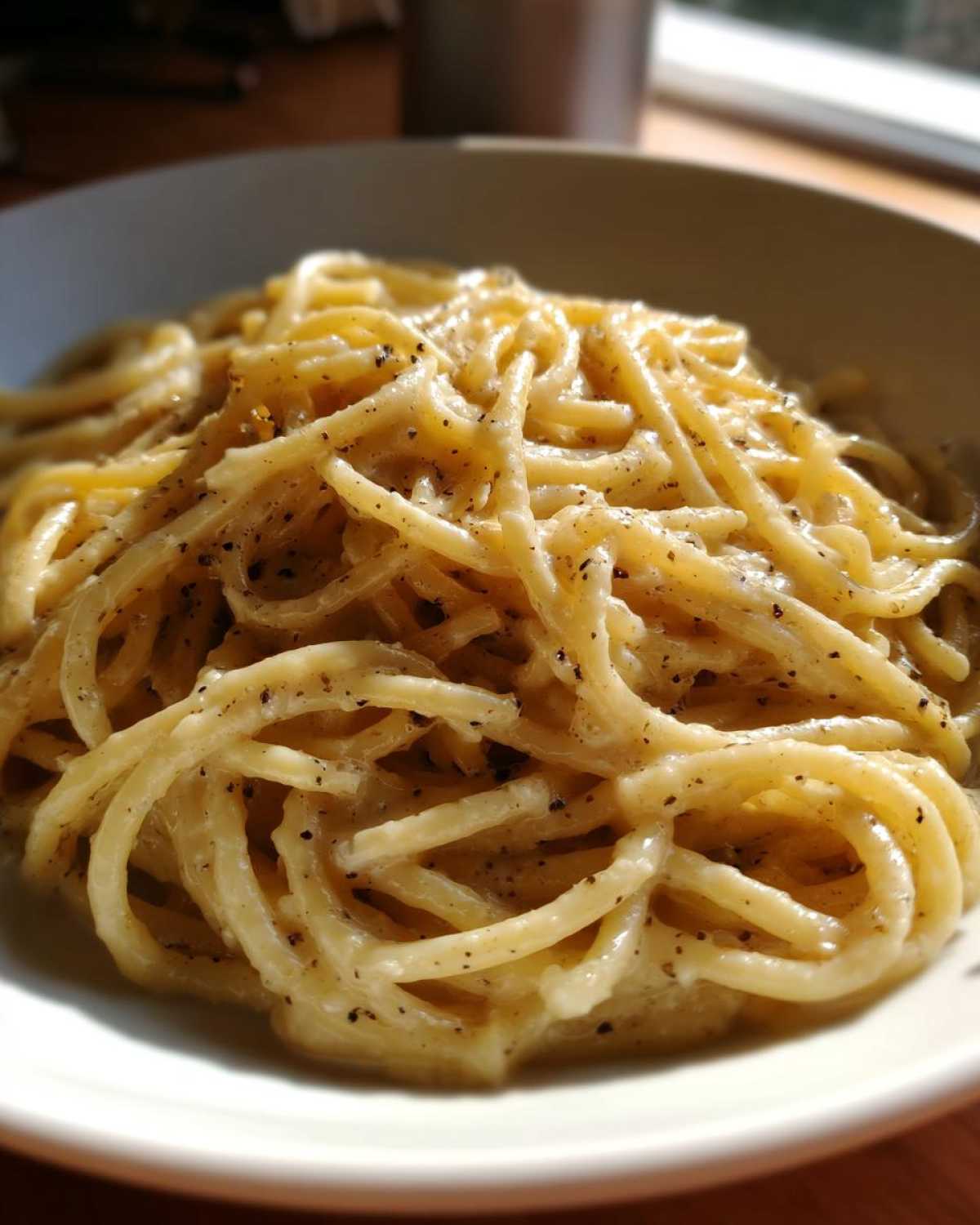 A close-up of a white bowl filled with creamy Spaghetti Cacio e Pepe, generously seasoned with black pepper.