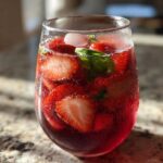 A close-up of a glass filled with Strawberry Basil Sangria, featuring sliced strawberries, basil leaves, ice, and bubbles.