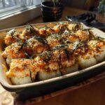 A close-up shot of a baked sushi casserole in a rectangular dish, topped with creamy sauce, breadcrumbs, and nori.