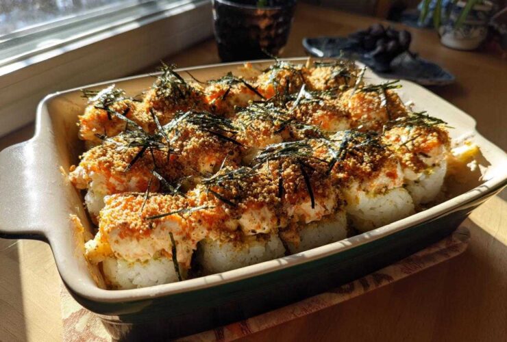 A close-up shot of a baked sushi casserole in a rectangular dish, topped with creamy sauce, breadcrumbs, and nori.