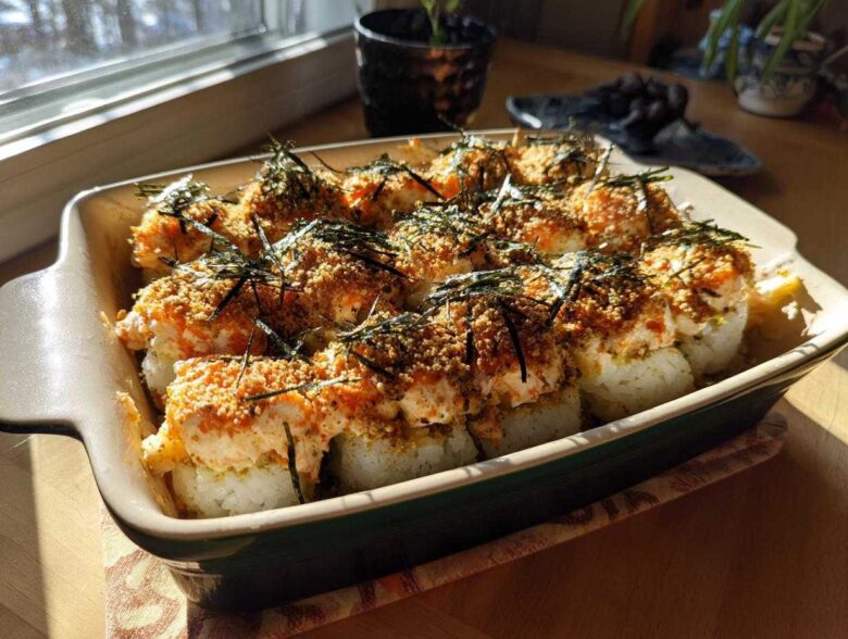 A close-up shot of a baked sushi casserole in a rectangular dish, topped with creamy sauce, breadcrumbs, and nori.
