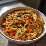 A close-up of a bowl filled with Thai Peanut Chicken Noodle Salad, featuring noodles, chicken strips, and shredded carrots, garnished with fresh herbs.