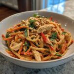 A bowl of Thai Peanut Chicken Noodle Salad with shredded chicken, carrots, red peppers, and noodles coated in peanut sauce.
