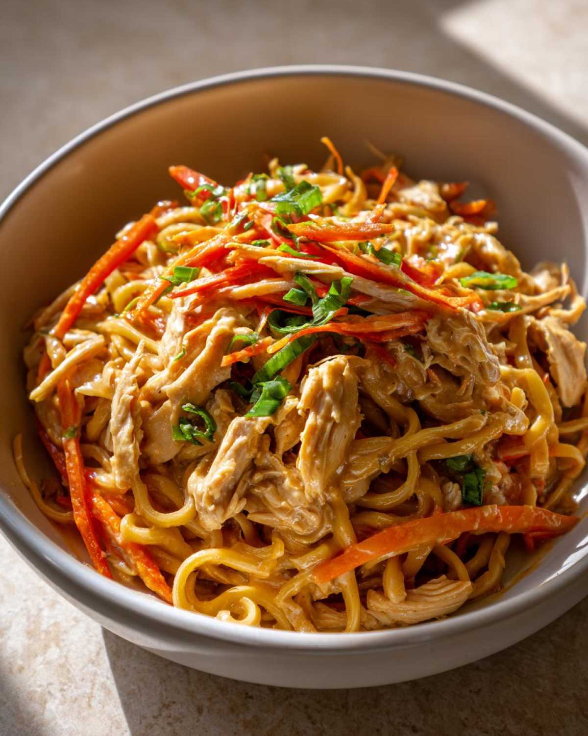 Close-up of a bowl of Thai Peanut Chicken Noodle Salad with shredded chicken, noodles, carrots, and green onions.