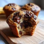 Close-up of a vegan chocolate chip muffin with a bite taken out, showing melted chocolate chips and a moist crumb.