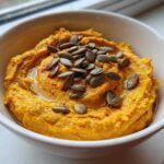 A bowl of creamy Vegan Pumpkin Hummus topped with pumpkin seeds and a sprinkle of paprika.