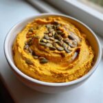 A close-up of creamy Vegan Pumpkin Hummus topped with pumpkin seeds and drizzled with oil in a white bowl.