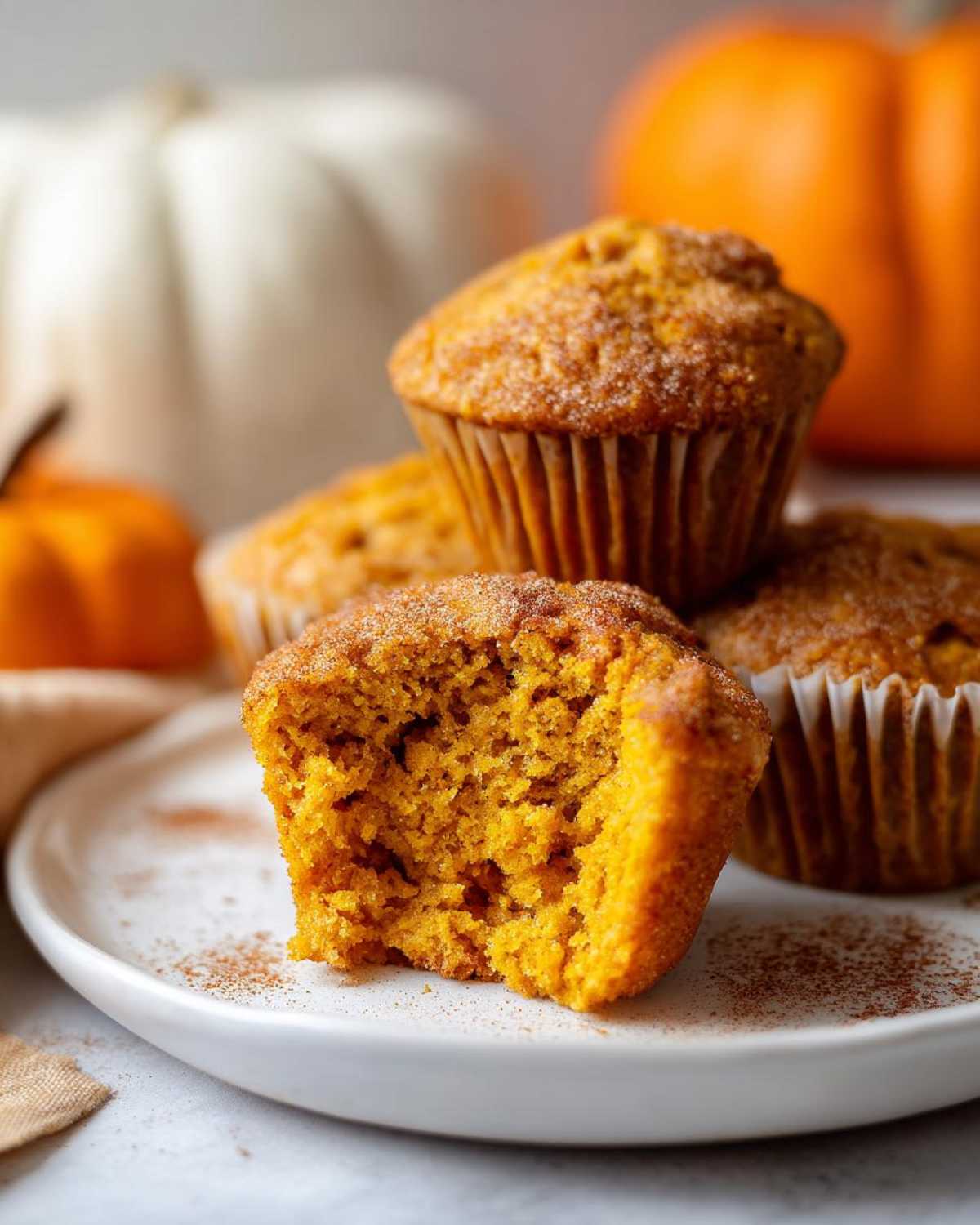 A close-up of a vegan pumpkin muffin, broken in half to reveal its moist interior, with more muffins and pumpkins in the background.