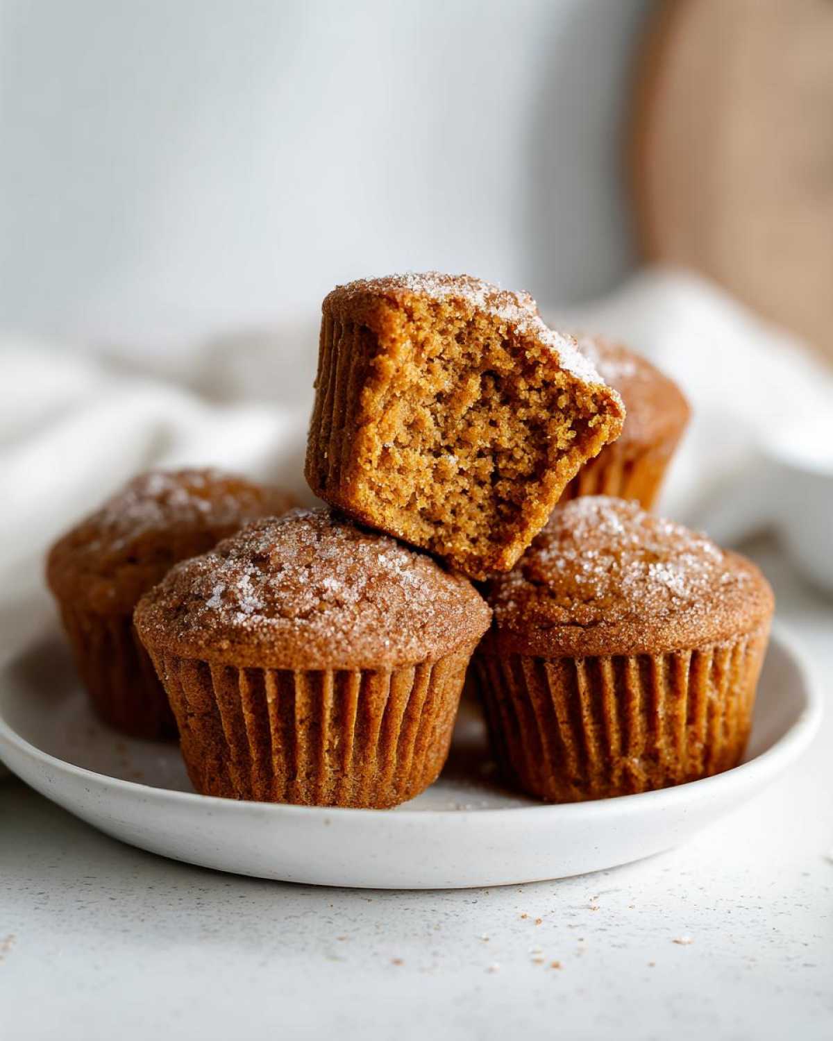 A stack of freshly baked vegan pumpkin muffins, with one muffin broken in half to show its moist interior and sugar-dusted topping.