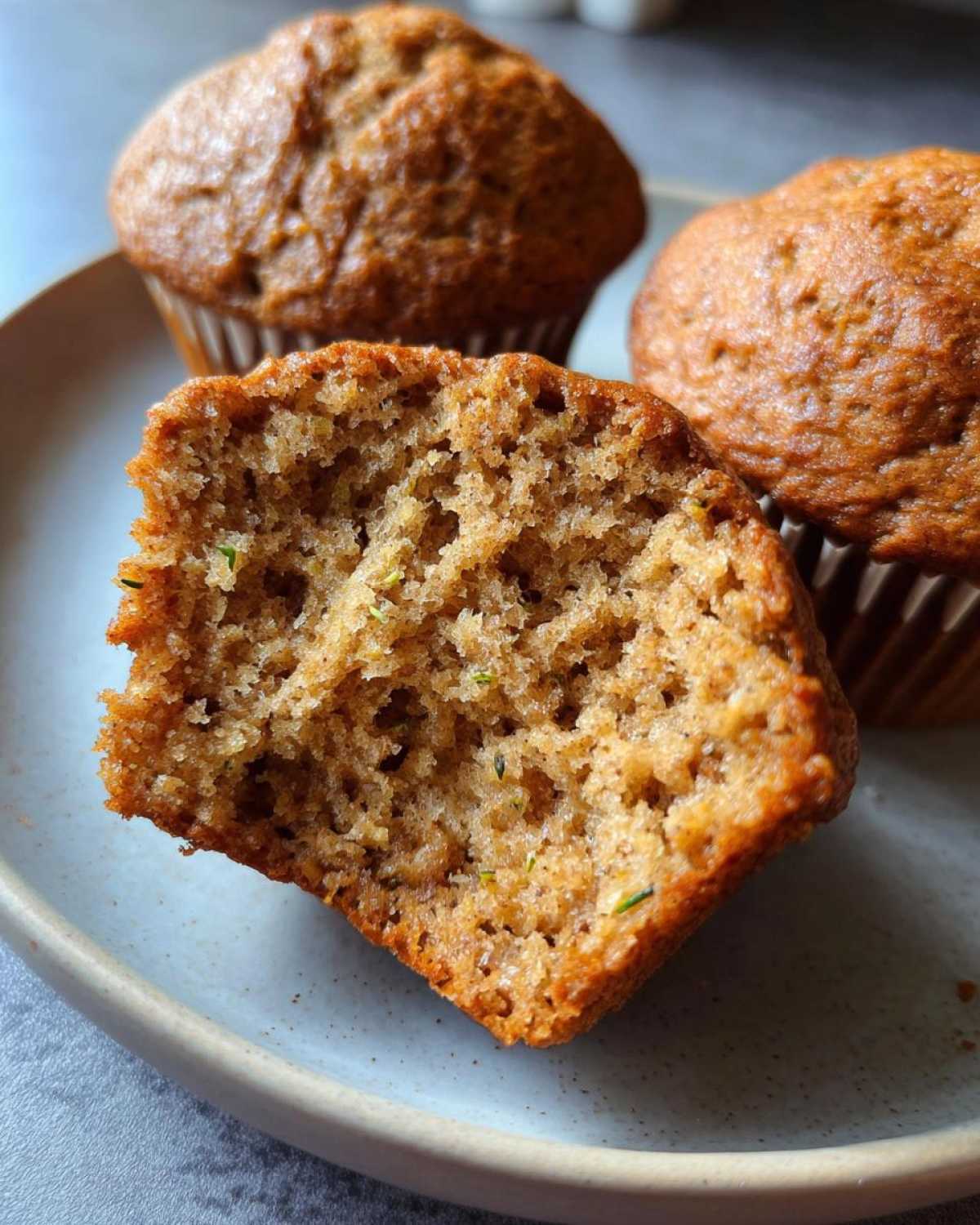 A close-up shot of a vegan zucchini muffin, broken in half to reveal its moist interior with visible shreds of zucchini.