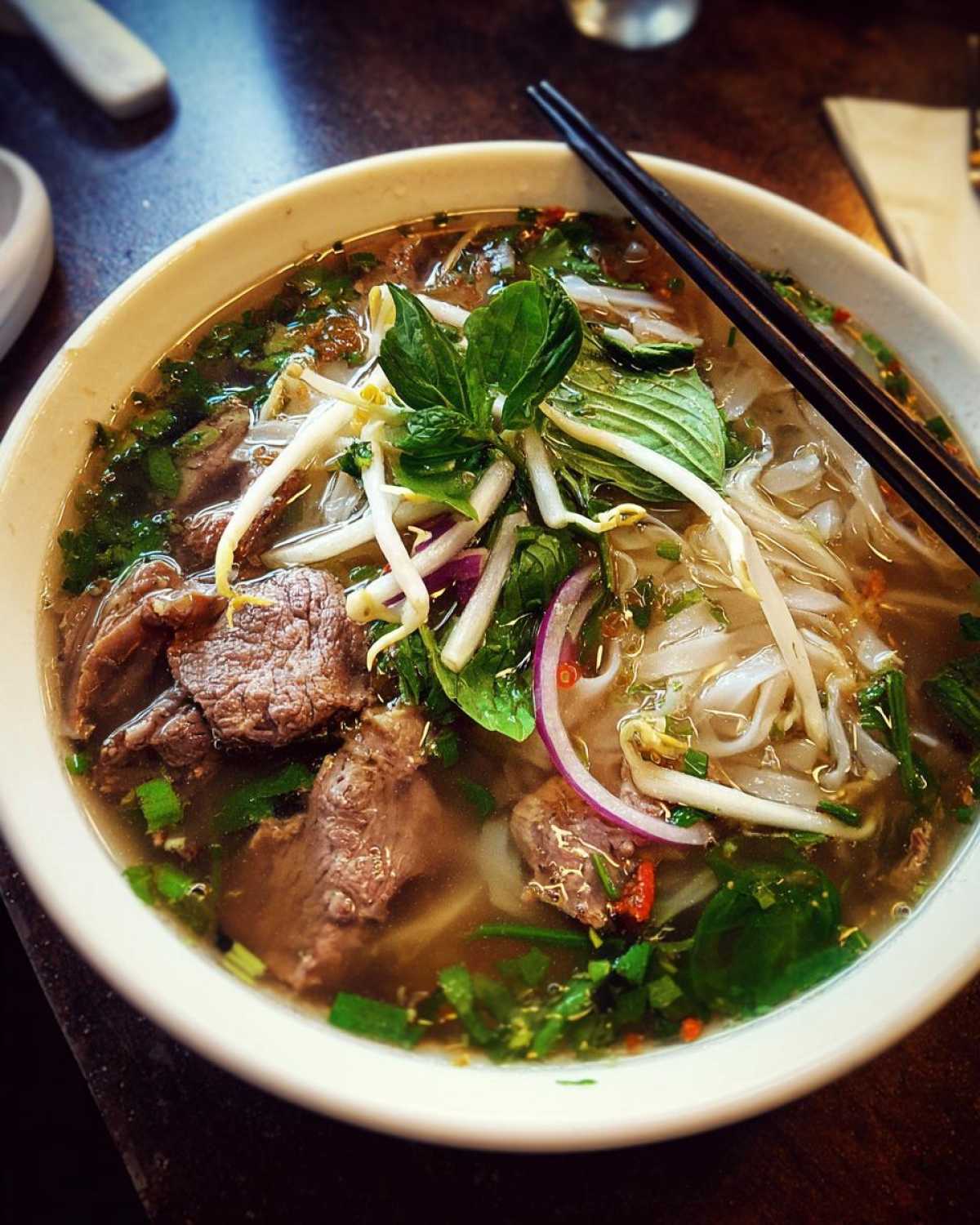 A close-up of a steaming bowl of Vietnamese Pho Soup, featuring tender beef slices, rice noodles, bean sprouts, fresh herbs, and red onion.