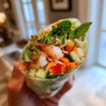 A hand holds a fresh Vietnamese Rice Paper Spring Roll filled with shrimp, cucumber, carrots, lettuce, and mint.