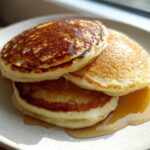 A stack of three golden brown almond flour pancakes drizzled with maple syrup on a speckled white plate.