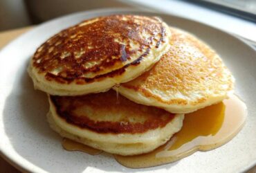 A stack of three golden brown almond flour pancakes drizzled with maple syrup on a speckled white plate.